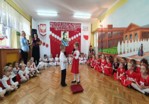 Chłopiec w białej koszuli i czarnych spodniach oraz dziewczynka w białej bluzce i czerwonej sukience na środku sali recytują wiersz.